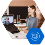 Two colleagues in an office share a video call; a smiling woman is at a laptop while another person sits nearby, with a blue icon overlay in the corner.