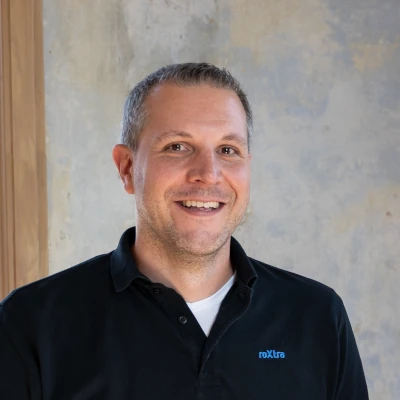 Smiling man in a black polo shirt with a small blue logo on the chest, standing indoors against a light, textured wall.